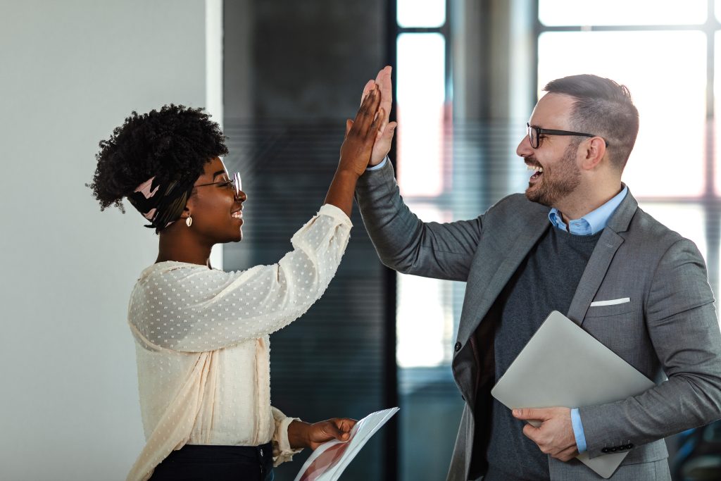 Coworkers celebrating achievement at office. Celebrating success. Happy young man standing in office and giving high five to his colleagues. Two business people high-five. Job well done